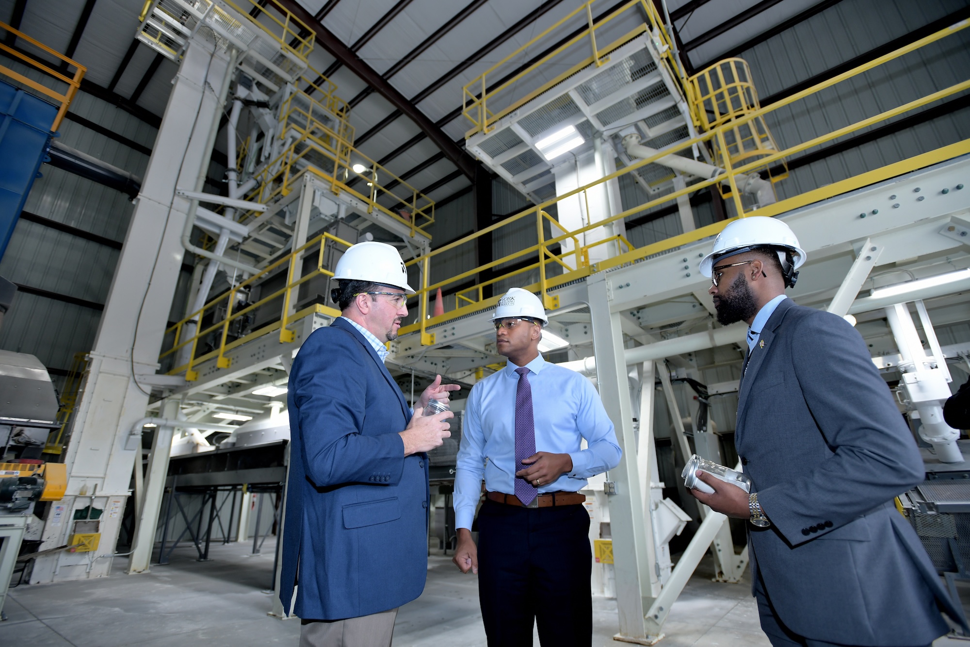 Three men in suits and hard hats holding jars with cement powder talking inside a manufacturing building